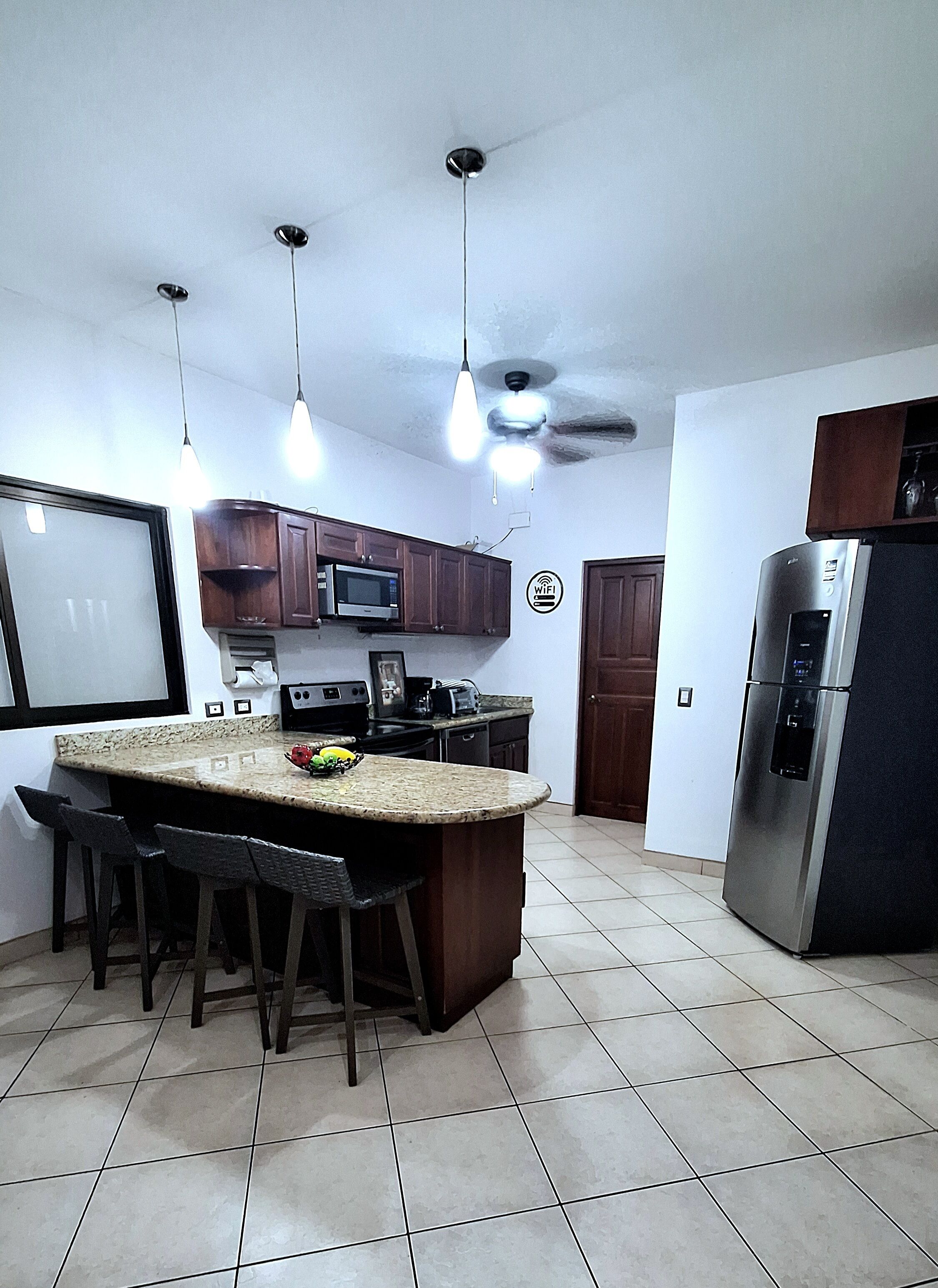 A kitchen with a central island, bar stools, a microwave, a refrigerator, pendant lights, and a ceiling fan.