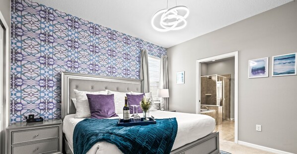 A modern bedroom with a patterned wallpaper, a large bed with a headboard, a nightstand, a tray with a bottle and glasses, and a bathroom visible through an open door. - Game On! Pool, Game Room & Themes | Near Disney (Kissimmee)