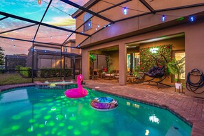 An outdoor swimming pool with inflatable flamingos and a ring, surrounded by a covered patio with string lights. - Game On! Pool, Game Room & Themes | Near Disney (Kissimmee)