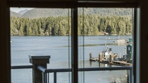 A room with a view of a lake, a chair, and a small table. - Island Vista - Waterfront Condo (Tofino)