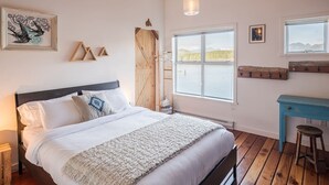 A bedroom with a wooden floor, a bed with white bedding, a wooden desk, and a window with a view of the outdoors. - Island Vista - Waterfront Condo (Tofino)