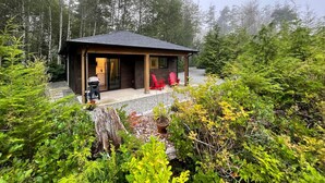 A cabin with a wooden exterior, a porch with red chairs, and a gravel driveway surrounded by dense forest. - Tidal Retreat Cabin in Tofino (Tofino)