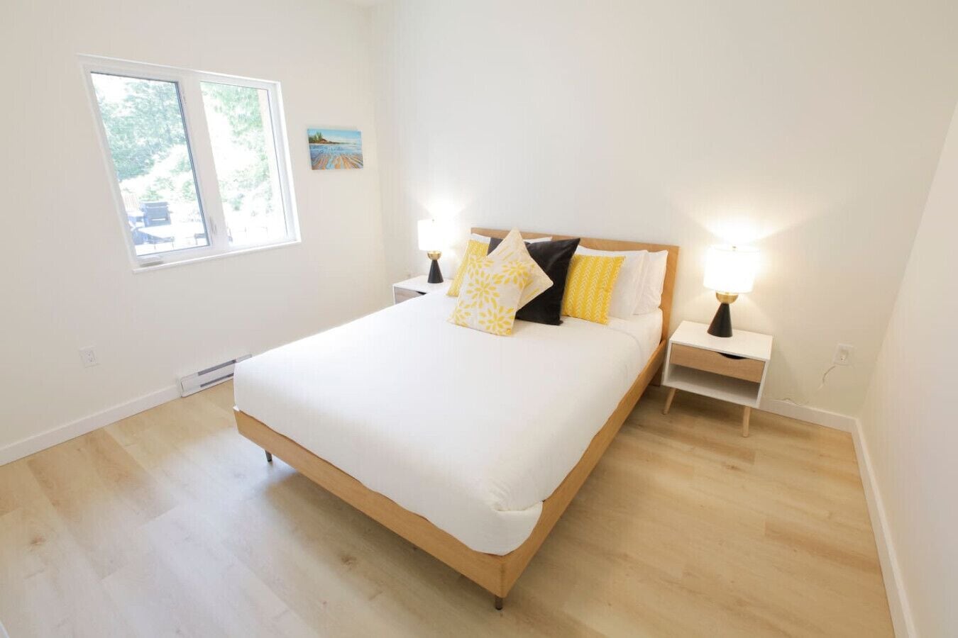 Un dormitorio con una cama grande, dos mesitas de noche con lámparas, una ventana y un cuadro pequeño en la pared.