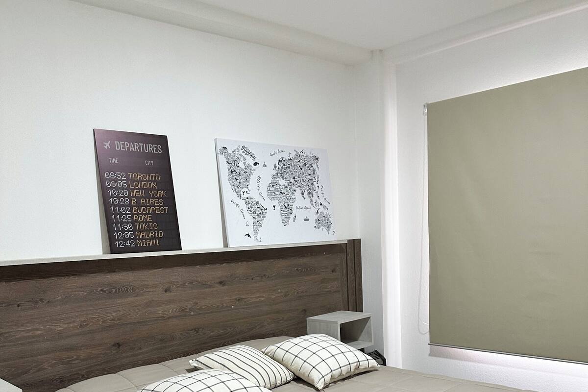 A neatly arranged hotel room with a bed, pillows, a nightstand, a wall-mounted departure board, and a world map.
