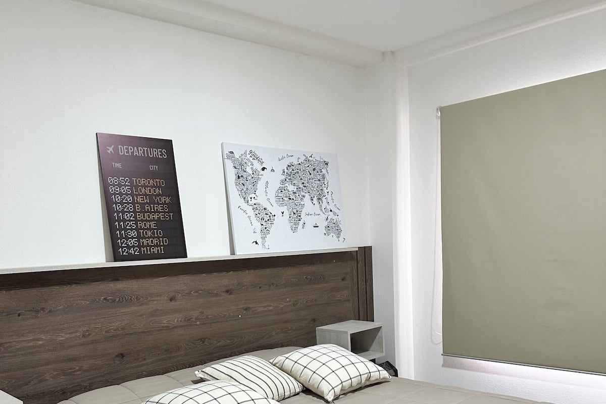 A neatly arranged hotel room with a bed, pillows, a nightstand, a wall-mounted departure board, and a world map.