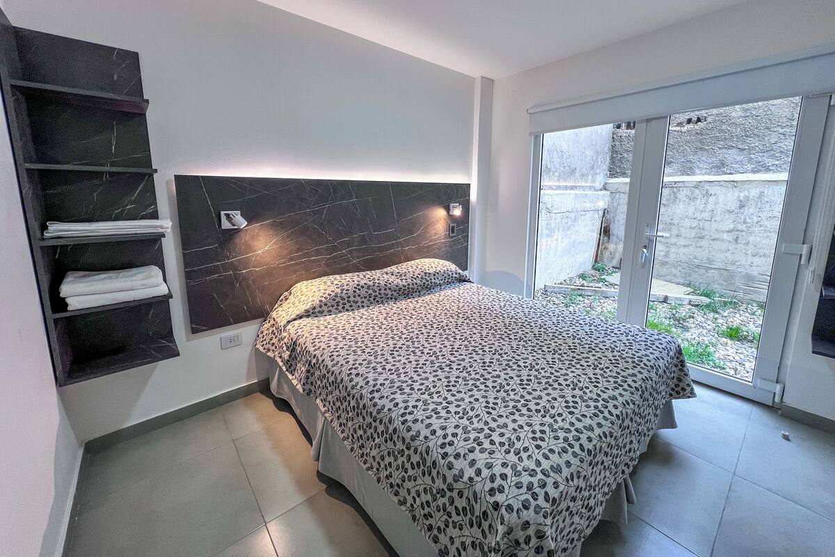 A bedroom with a bed, a black marble headboard, a shelf with towels, and a view through a glass door.