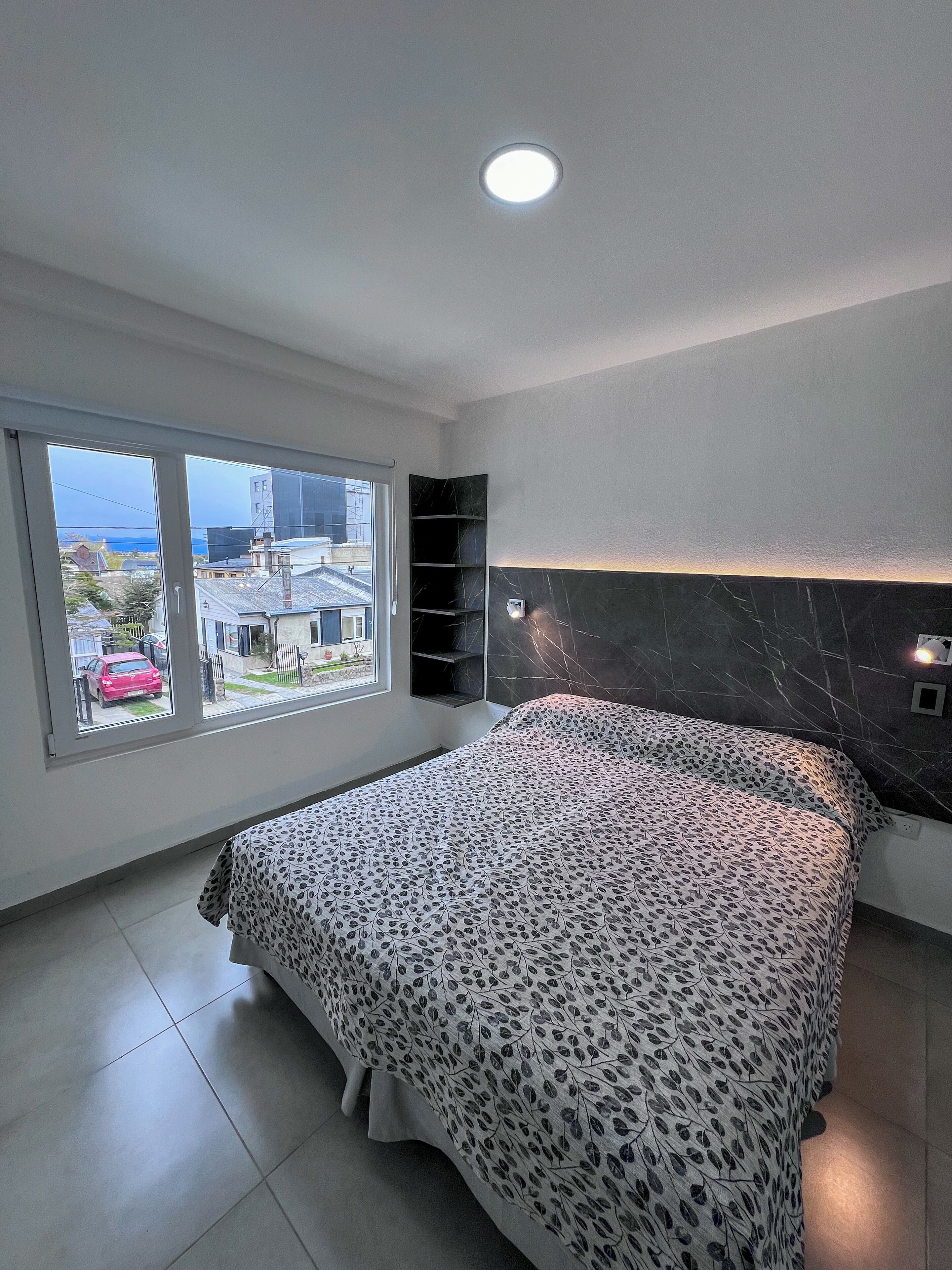A bedroom with a large bed, a built-in shelf, a window with a view of the outside, and a ceiling light.