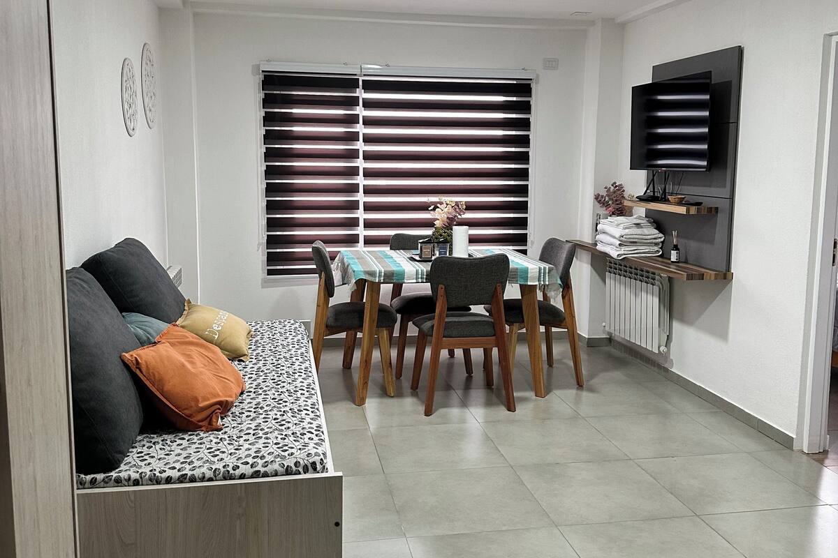 A modern living room with a sofa, dining table, chairs, a TV mounted on the wall, and a window with blinds.