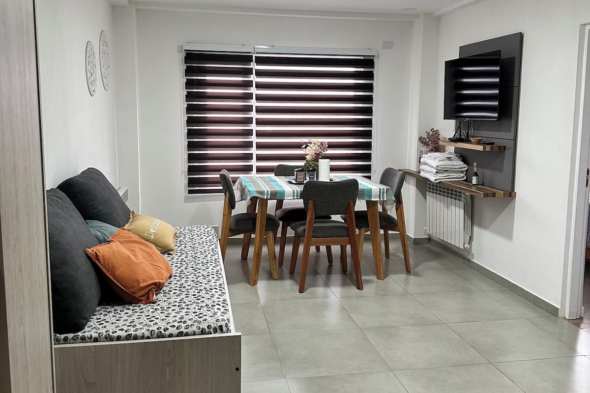 A modern living room with a sofa, dining table, chairs, a TV mounted on the wall, and a window with blinds.