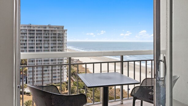 Un balcon avec une table et des chaises, donnant sur une plage et un immeuble de grande hauteur.