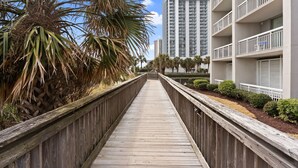 Une promenade en bois mène à un bâtiment moderne doté de balcons, sous un ciel dégagé.