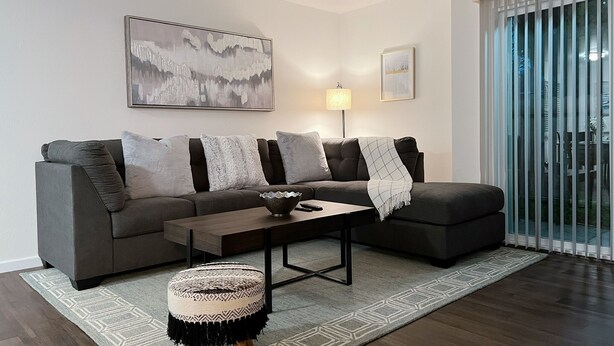 A modern living room with a grey sectional sofa, a round ottoman, a coffee table, and a floor lamp. There is a large window with blinds and a painting on the wall. - Super Convenient, Cozy 2B1b Apartment, Berkeley (Berkeley)