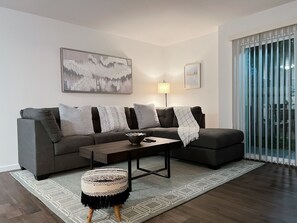 A modern living room with a grey sectional sofa, a round ottoman, a coffee table, and a floor lamp. There is a large window with blinds and a painting on the wall. - Super Convenient, Cozy 2B1b Apartment, Berkeley (Berkeley)