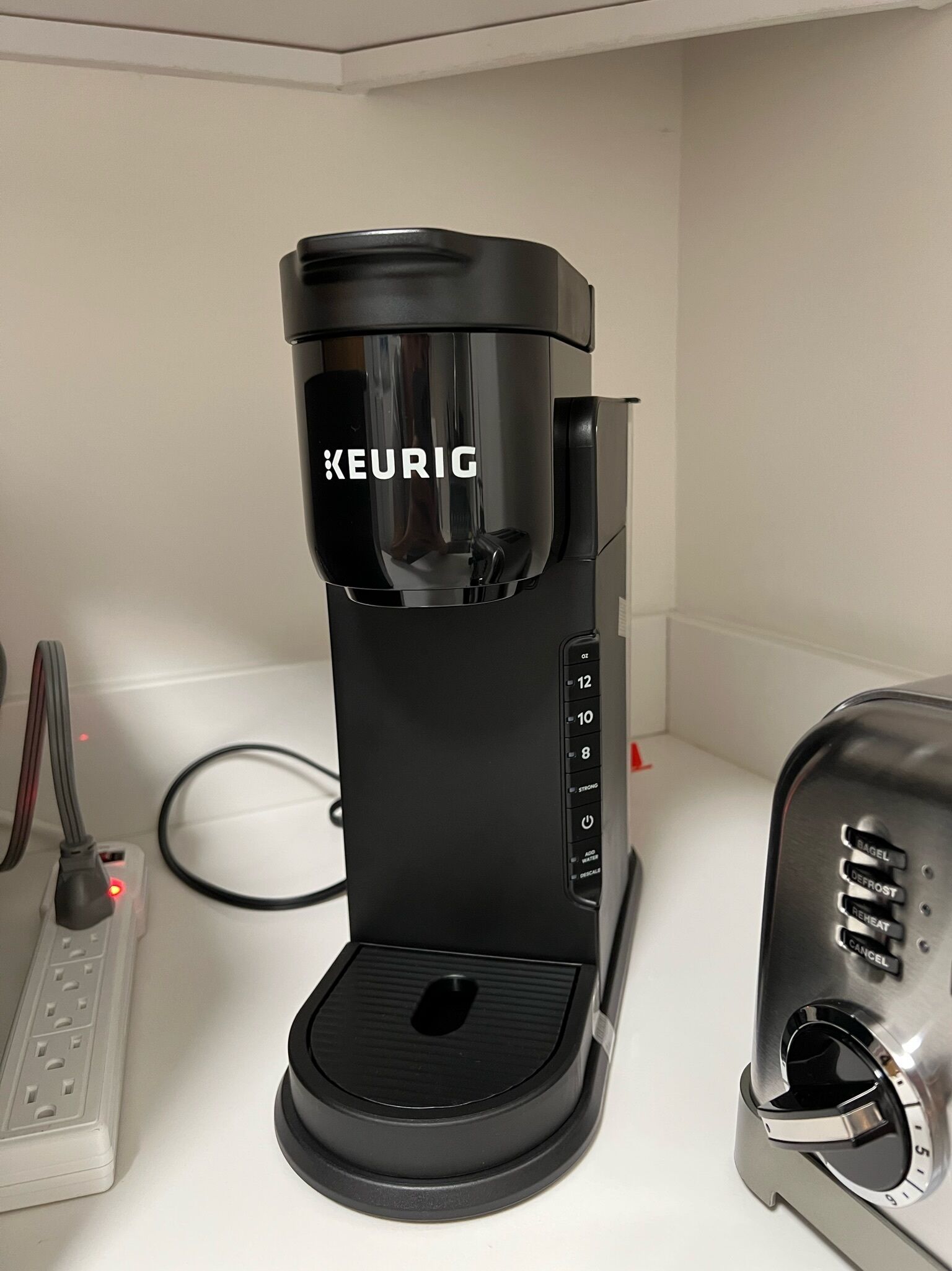 A black Keurig coffee maker on a white countertop with a silver toaster and a power strip with a red light.