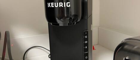 A black Keurig coffee maker on a white countertop with a silver toaster and a power strip with a red light.
