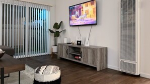 A modern living room with a flat-screen TV mounted on the wall, a wooden entertainment unit, a large potted plant, and a sliding glass door leading to an outdoor area. - Super Convenient, Cozy 2B1b Apartment, Berkeley (Berkeley)