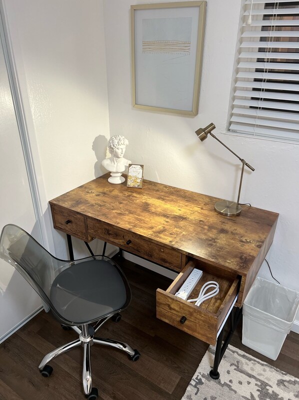 Un bureau en bois avec un tiroir, une chaise de bureau en verre, une sculpture en buste, un tableau encadré et une lampe sur pied.