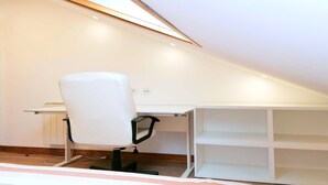 Un pequeño y luminoso cuarto ático con un escritorio y silla blancos, techo inclinado y estanterías empotradas.