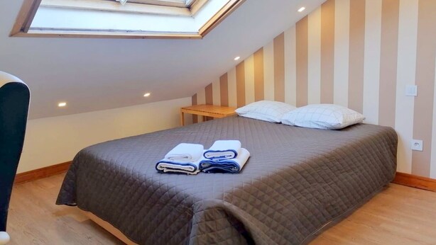 Una habitación con tragaluz, una cama con colcha acolchada, dos almohadas y una pequeña mesa de madera.