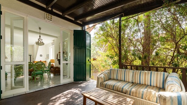 An outdoor deck with a wicker sofa and chairs, a wooden table, and a glass door leading to an interior space with a lamp and a chair.