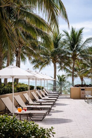 A row of sun loungers with umbrellas, a poolside table with drinks, and palm trees in the background. - 1 bed condo at the Palm Beach Marriott with Hotel Amenities and views RENOVATED (Riviera Beach)