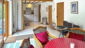 Een ruime kamer met een hoge plafond, een eethoek met rieten meubels, en een keuken die zichtbaar is door een open deur.