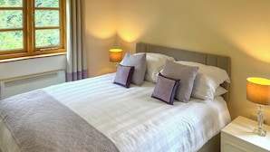 Een opgemaakt bed met een beddekleed met patroon en kussens, een bedlampje en een raam met gordijnen.