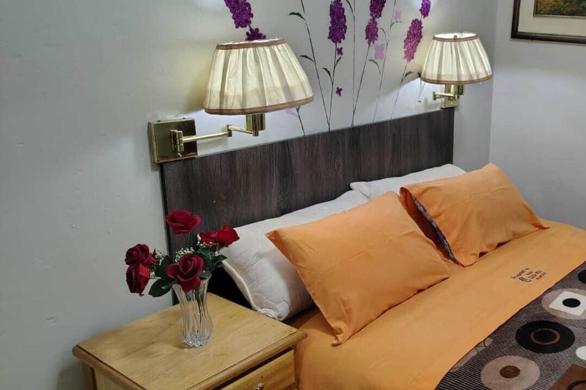 Una cama bien hecha con una colcha naranja y dos almohadas, un jarrón con rosas rojas sobre una mesita de noche y dos lámparas de pared.