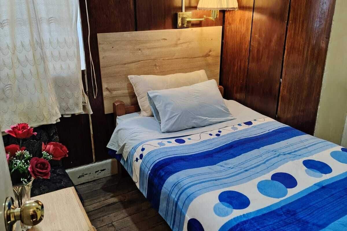 Un dormitorio con cabecera de madera, cama con colcha azul y blanca estampada, lámpara de noche y mesita de noche de madera con un jarrón de flores rojas.