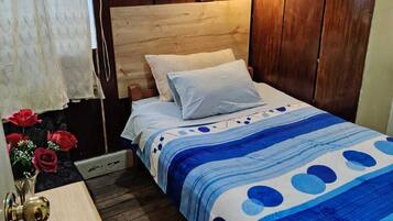 Een slaapkamer met een houten hoofdbord, een bed met een blauw-witte bedspreidek, een bedlamp en een klein houten nachtkastje met een vaas rode bloemen.