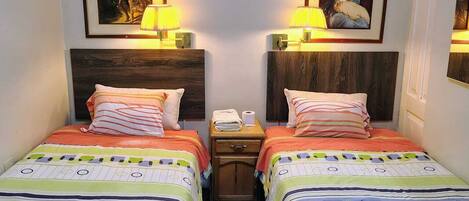 A hotel room with two beds, each with a colorful striped bedspread. There are framed pictures on the walls above the beds, and two bedside lamps.