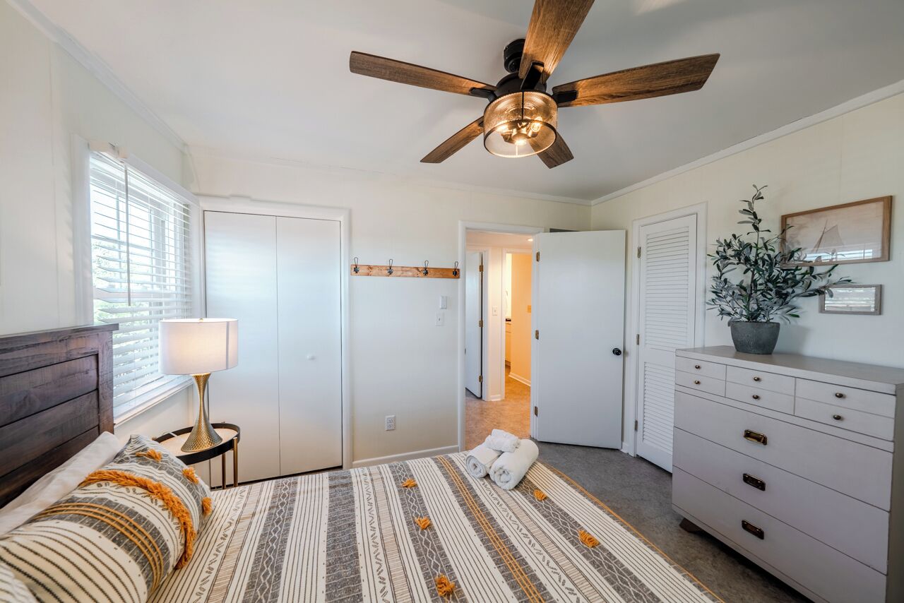 A bedroom with a bed, a ceiling fan, a nightstand, a dresser, and a potted plant.