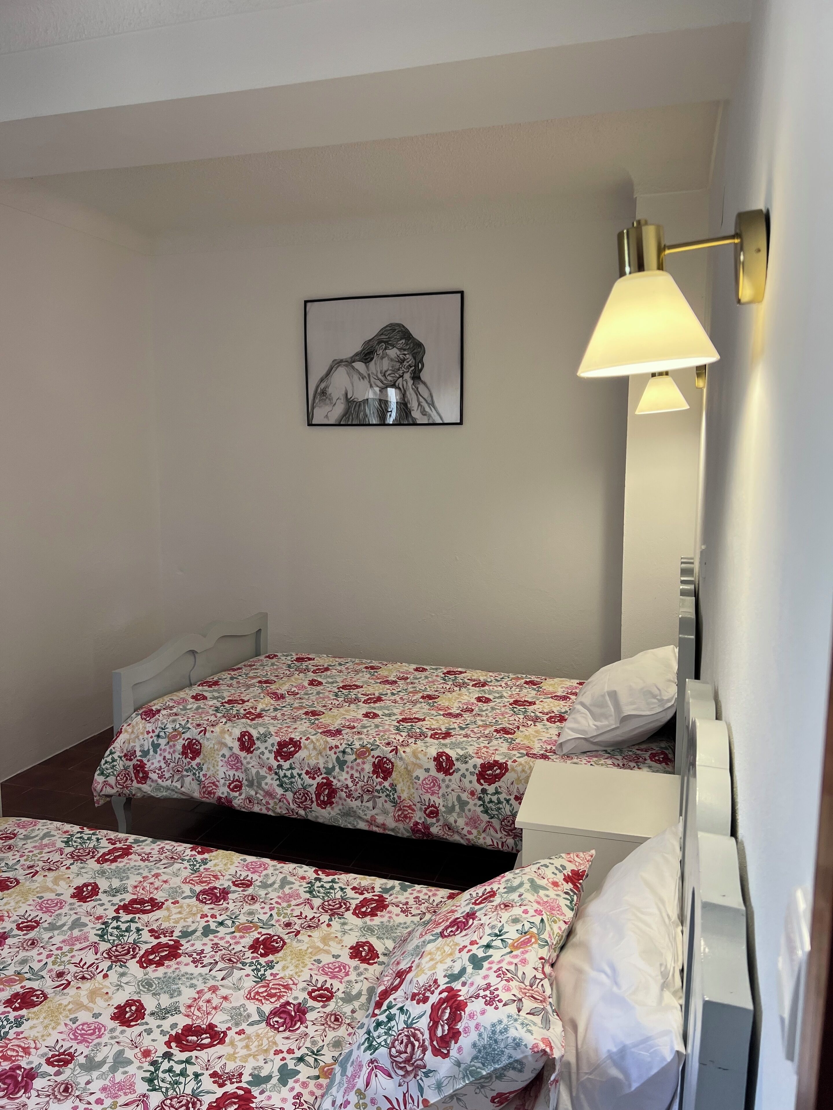 A hotel room with two single beds, floral bedding, a framed artwork on the wall, and a wall-mounted lamp.