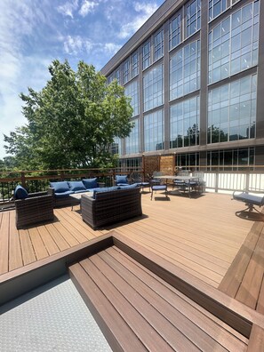 A wooden deck with outdoor furniture, a large building with reflective windows, and a tree in the background.