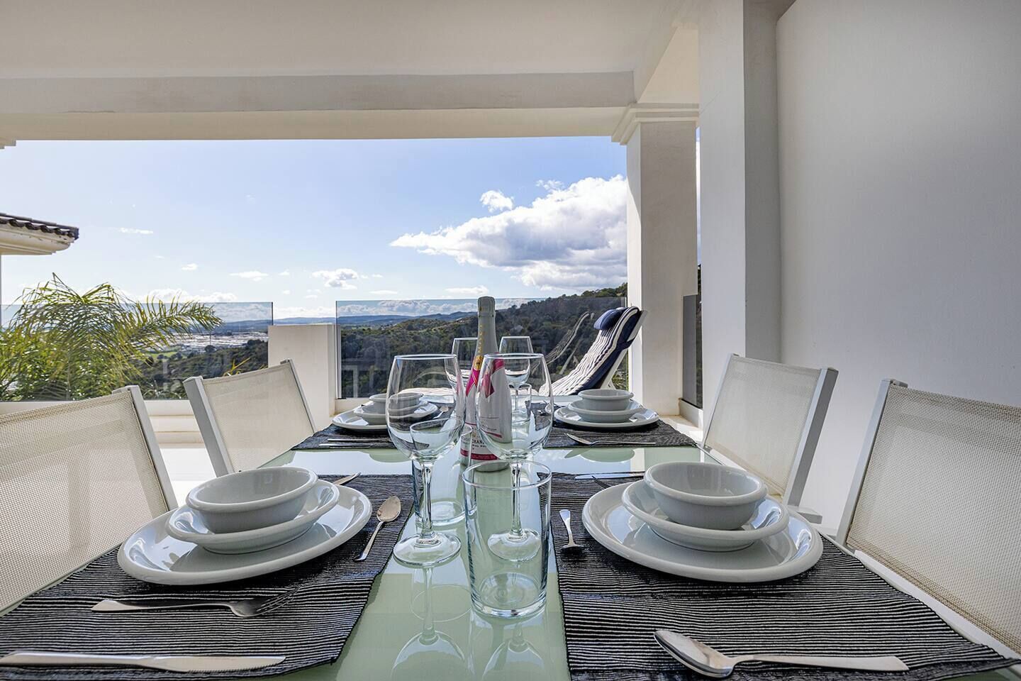 Une table à manger dressée pour six personnes, avec une vue magnifique, des chaises blanches et une bouteille de champagne.