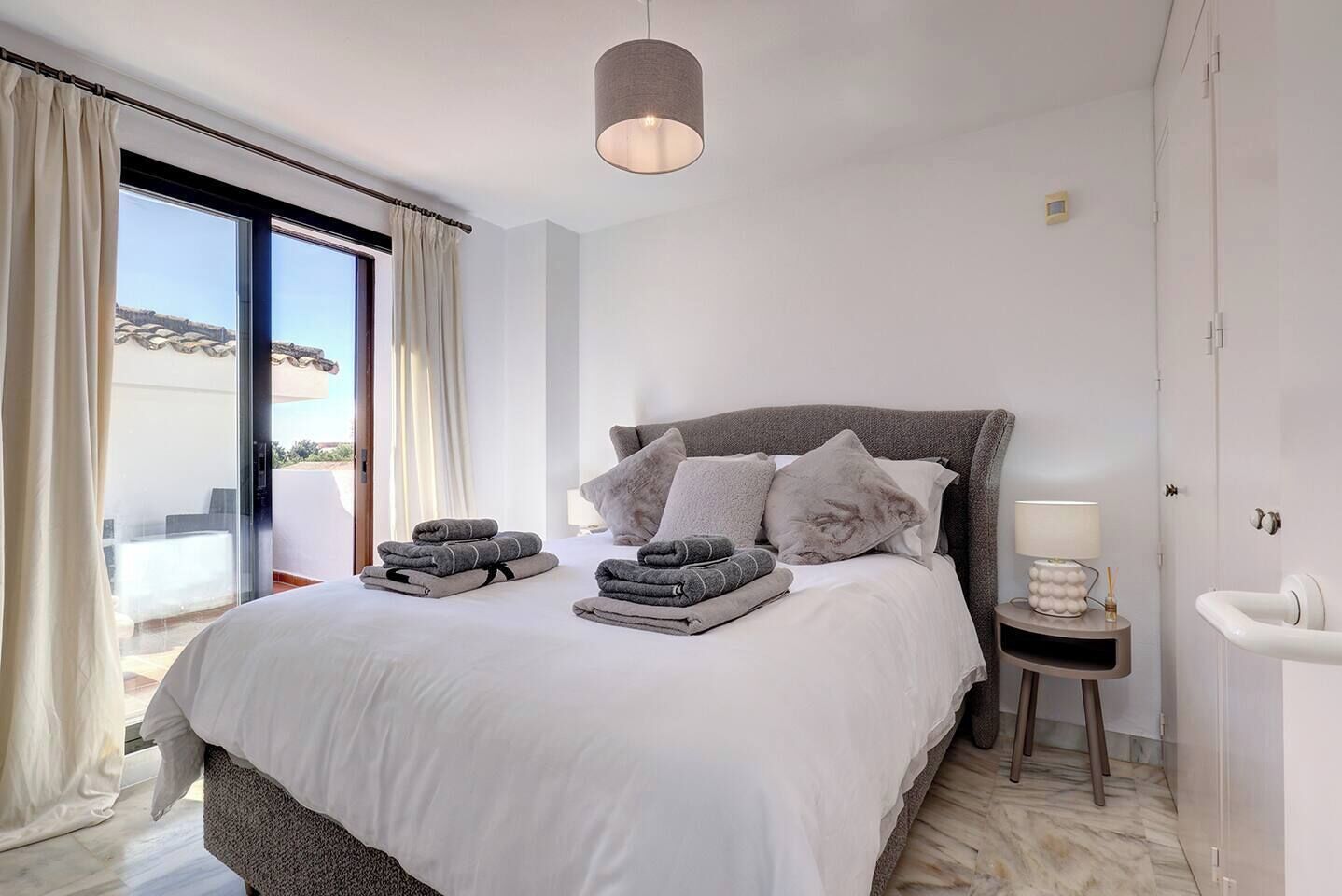 A modern bedroom with a large bed, a bedside table with a lamp, and a view of the outdoors.