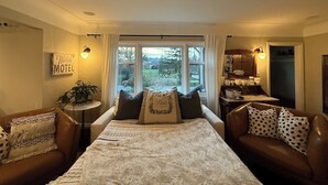 A cozy room with a large bed, two armchairs, a small table, and a view of the outdoors through large windows. - Couple's Oasis at Horner Park, Welch Homes & Hospitality, pet friendly property. (Saanich)