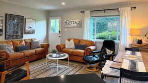 A cozy living room with two brown leather sofas, a coffee table, and a dining area with a wooden table and chairs. - Family Cottage at Horner Park, Welch Homes & Hospitality, pet friendly property. (Saanich)
