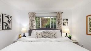 A bedroom with a large bed, two bedside tables with lamps, a window with curtains, and framed artwork on the walls. - Family Cottage at Horner Park, Welch Homes & Hospitality, pet friendly property. (Saanich)