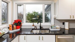 A kitchen with white cabinets, a black countertop, a sink, and a window with a view of trees. - Family Cottage at Horner Park, Welch Homes & Hospitality, pet friendly property. (Saanich)