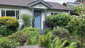A house with a blue front door, surrounded by lush greenery and a small garden. - Family Cottage at Horner Park, Welch Homes & Hospitality, pet friendly property. (Saanich)