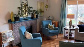 Ein Wohnzimmer mit Kamin, blauen Sesseln, einem grünen Sessel, einem kleinen Tisch mit Lampe und Blick ins Freie durch ein Fenster.