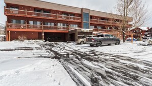 Um hotel com vários andares, fachada vermelha e varandas. Vários veículos estacionados em frente, e chão coberto de neve com marcas de pneus visíveis.