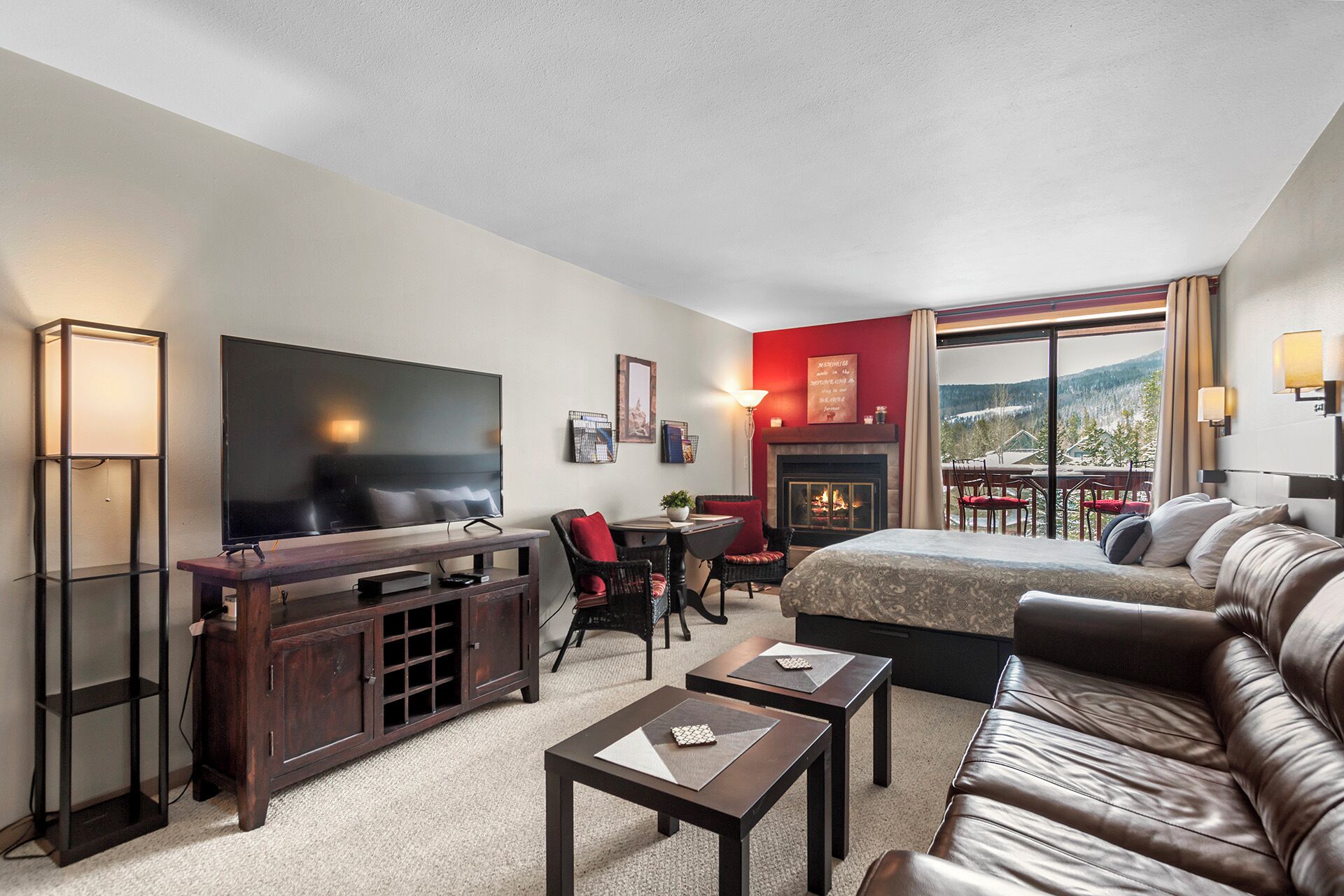 A modern hotel room with a large flat-screen TV, a fireplace, a sofa, and a dining area with a view of the mountains.