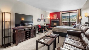 Quarto de hotel moderno com uma televisão de tela grande, lareira, sofá e área de jantar com vista para as montanhas.
