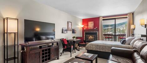 A modern hotel room with a large flat-screen TV, a fireplace, a sofa, and a dining area with a view of the mountains.