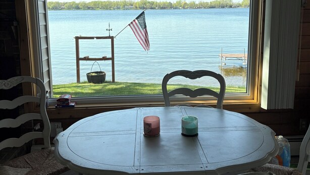 A dining area with a table and chairs, a ceiling fan, a view of a lake with an American flag, and a sign that says "HAVE FAITH". - Finch House on Lake Osakis: Charming 2-bedroom cabin (Osakis)