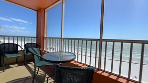 A balcony with a round table and two chairs, overlooking a beach with a clear sky.