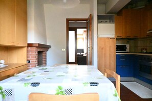 Un comedor con una mesa cubierta por un mantel estampado, sillas de madera, una chimenea de ladrillo y vista a una cocina con armarios azules y un microondas.