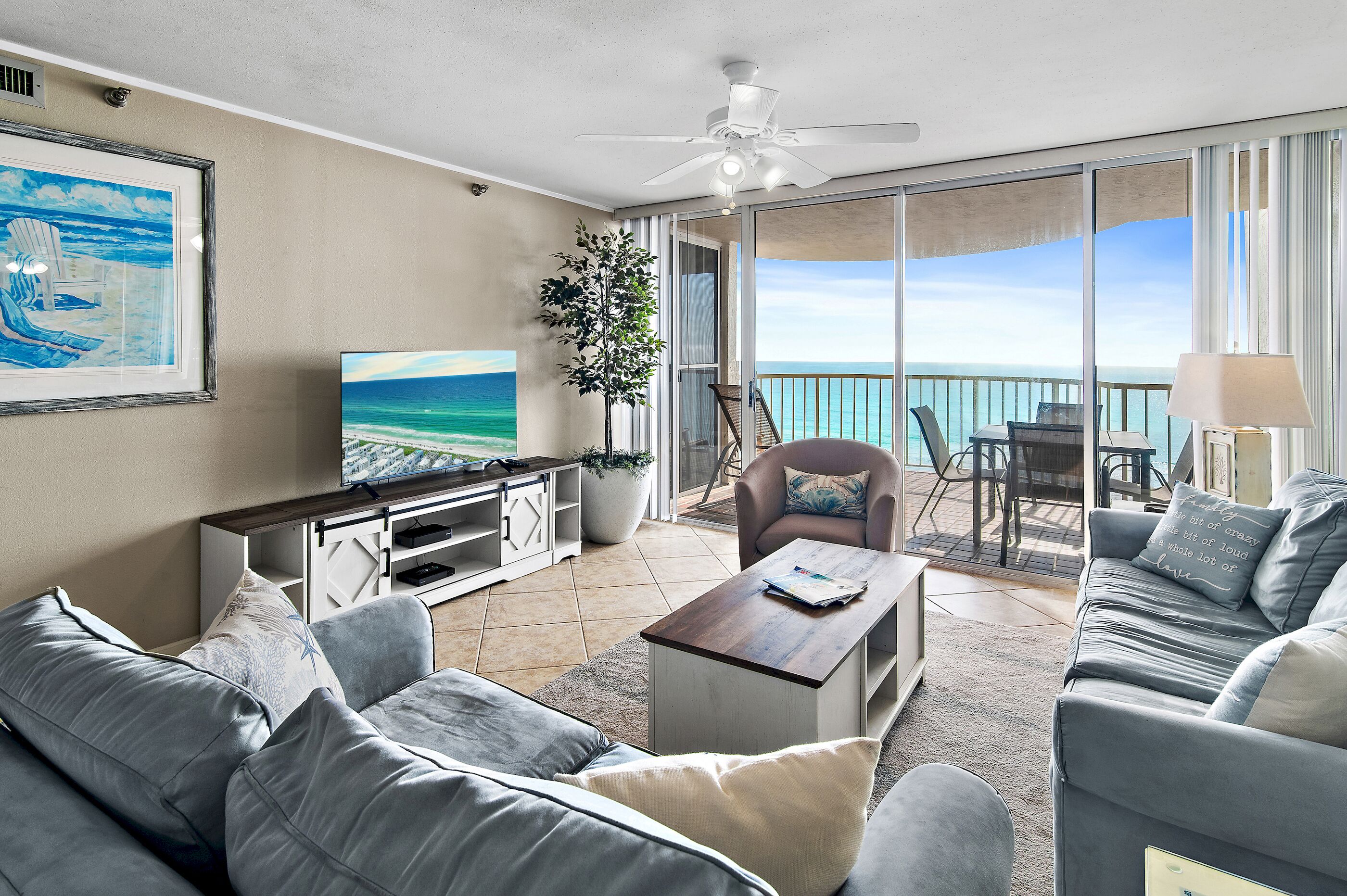 A modern living room with a large TV, comfortable seating, a ceiling fan, and a balcony with a view of the ocean.